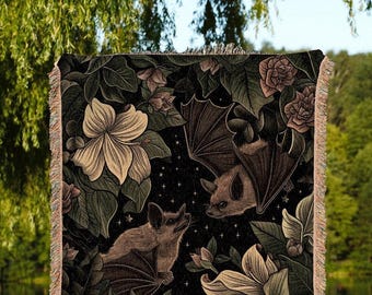 Puede incluir: Una manta tejida rectangular con fondo oscuro, dos murciélagos, grandes flores pálidas y hojas verdes. El diseño incluye pequeñas estrellas blancas dispersas. La manta tiene un borde con flecos en los cuatro lados.