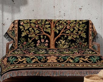 May include: A black throw blanket with a tree design draped over a wooden sofa. The tree has brown branches, green leaves, and pink flowers. The blanket has a fringe along the edges. A floor lamp and a woven basket are also in the image. The text "INK & SHADOW Original Fine Art Throws" is at the bottom.