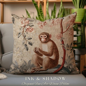 Op de afbeelding: Een decoratief kussen met een geweven ontwerp van een aap die in een boom zit met roze bloemen. Het kussen is beige met bruine, groene en roze accenten. De tekst "INK & SHADOW Original Fine Art Woven Pillow" is onderaan het kussen gedrukt.