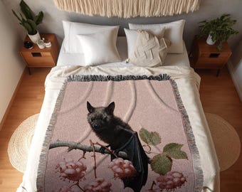 Puede incluir: Una manta tejida que presenta un murciélago marrón oscuro posado en una rama con flores rosas. La manta tiene un borde con flecos y muestra el texto INK & SHADOW Original Fine Art Throws en la parte inferior central sobre un fondo rosa.