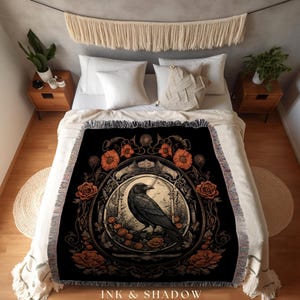 May include: Black throw blanket featuring a detailed illustration of a raven within a floral frame. The design includes orange flowers and roses. The text "INK & SHADOW Original Fine Art Throws" is at the bottom.