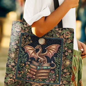 May include: A tote bag featuring a dragon perched atop a stack of books under a full moon and starry sky. The bag has a floral border and the text "INK & SHADOW Woven Fine Art Tote". The bag is carried by a person.
