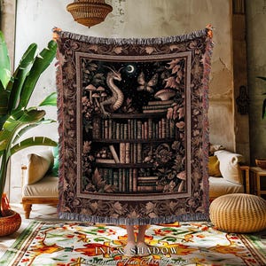 May include: A woven throw blanket featuring a dark, detailed illustration of a dragon perched atop a bookshelf filled with books, mushrooms, and a butterfly under a crescent moon. The blanket has a decorative floral border and fringed edges.
