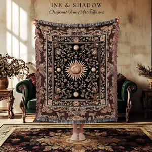 May include: A woven throw blanket featuring a central sun and moon motif surrounded by floral patterns in shades of brown, black, and cream. The rectangular textile has a fringed border and displays the text INK & SHADOW Original Fine Art Throws.