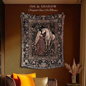 May include: A woven throw blanket featuring a dark, intricate scene of a person with long hair standing beside a white unicorn in a forest. The design includes a floral border and fringed edges, with the text INK & SHADOW Original Fine Art Throws.