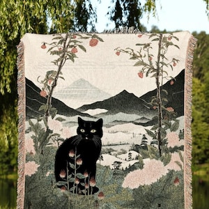 May include: A woven throw blanket featuring a black cat in a landscape scene with mountains, trees, and flowers. The blanket has a fringed edge and the text "JOY OF PLANTS Original Fine Art Throws".