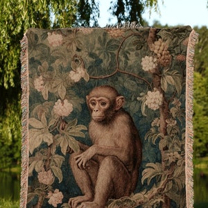 May include: A woven tapestry throw blanket featuring a detailed illustration of a monkey sitting in a tree with flowers and leaves. The throw blanket is a dark blue, green, and brown colour. The text "INK & SHADOW Original Fine Art Throws" is printed at the bottom of the image.
