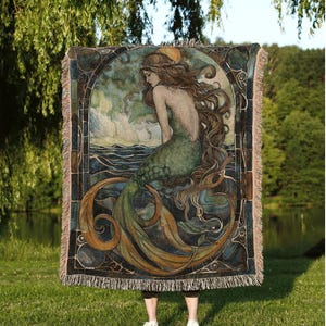 May include: A decorative throw blanket with a mermaid design in green, blue, and gold tones. The image shows a mermaid with long hair, near water. The blanket has a fringed edge and the text "INK & SHADOW Original Fine Art Throws".