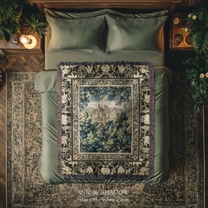 May include: A woven throw blanket featuring a central rectangular scene of a castle nestled within a dense forest. The border displays a repeating pattern of cream-coloured floral motifs against a dark background with fringed edges on all sides.