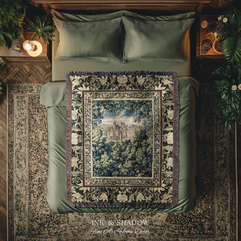 May include: A woven throw blanket featuring a central rectangular scene of a castle nestled within a dense forest. The border displays a repeating pattern of cream-coloured floral motifs against a dark background with fringed edges on all sides.