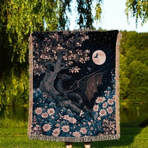 May include: A woven throw blanket featuring a fantasy scene with a black dragon, a full moon, and a tree with pink and white flowers. The blanket has a fringed edge and the text "INK & SHADOW Original Fine Art Throws" at the bottom.
