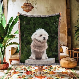 May include: A fringed throw blanket with a picture of a white dog wearing a sweater, set against a green background. The blanket is displayed in a room with a patterned rug and a woven lampshade. The text "INK & SHADOW Original Fine Art Prints" is visible.