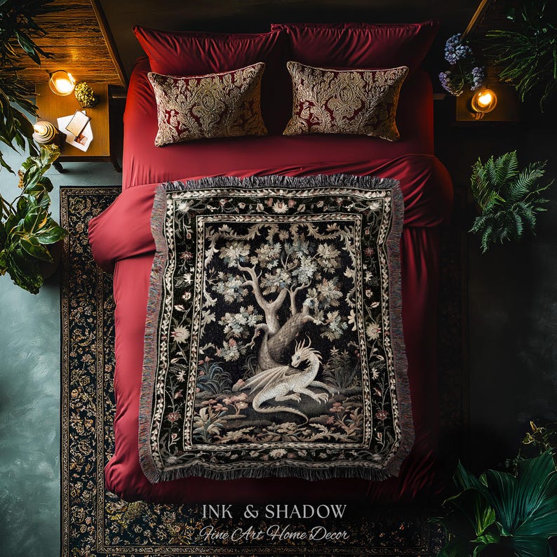 May include: A woven throw blanket featuring a detailed illustration of a white dragon resting beneath a large tree, set against a dark background with floral borders. The blanket is draped over a bed with deep red bedding and two patterned pillows.