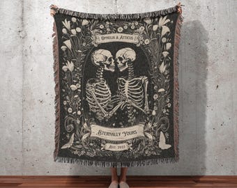 May include: A dark gray woven throw blanket featuring a romantic skeleton couple design. The blanket has a floral border and the text "Ophelia & Atticus" and "Eternally Yours Est. 2025". The bottom of the blanket reads "INK & SHADOW Original Fine Art Throws".