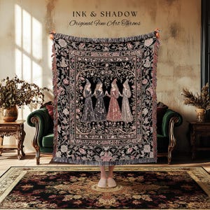 May include: A woven throw blanket featuring a central illustration of four figures in pointed hats standing in a circle, surrounded by a detailed floral border in black, pink, and cream tones. The text INK & SHADOW Original Fine Art Throws is visible.