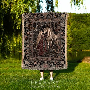 Puede incluir: Una manta de tapiz negra con una escena de bosque de una mujer y un unicornio. La manta tiene un borde con flecos y un borde floral. El texto "INK & SHADOW Original Fine Art Throws" está en la parte inferior.
