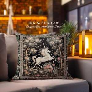 Może przedstawiać: Czarno-biała poduszka tkana z białym jednorożcem w leśnym otoczeniu z r&oacute;żowymi kwiatami. Poduszka ma napis "INK & SHADOW Original Fine Art Woven Pillow".