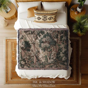 May include: A woven throw blanket with a forest scene in shades of green, brown, and gray is draped over a bed. The bed has white linens, a patterned pillow, and a wooden frame. The blanket has a fringed edge. The text "INK & SHADOW Fine Art Home Decor" is at the bottom.