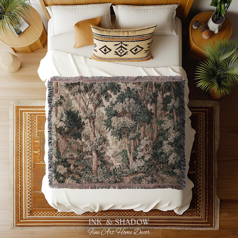 May include: A woven throw blanket with a forest scene in shades of green, brown, and gray is draped over a bed. The bed has white linens, a patterned pillow, and a wooden frame. The blanket has a fringed edge. The text "INK & SHADOW Fine Art Home Decor" is at the bottom.