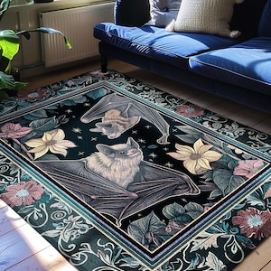 Puede incluir: Una alfombra rectangular con fondo negro que presenta dos murci&eacute;lagos, uno colgado y otro erguido, rodeados de flores y hojas estilizadas en tonos rosa, amarillo y azul verdoso. El texto dice INK & SHADOW Original Fine Art Rugs.