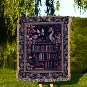 May include: A tapestry throw blanket depicting a fantasy scene with a dragon, butterfly, and mushrooms in front of a bookshelf. The throw has a dark background with a decorative border and fringe. The text "INK & SHADOW Original Fine Art Throws" is at the bottom.