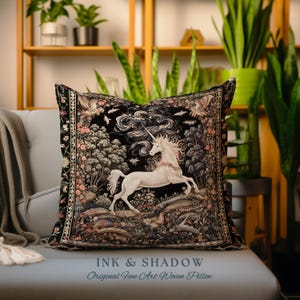 K&ouml;nnte beinhalten: Ein schwarzes und wei&szlig;es Wandteppichkissen mit einem Einhorn, das durch einen Wald rennt. Das Kissen hat einen dunklen Hintergrund mit filigranen Details und einem floralen Rand. Der Text "INK & SHADOW Original Fine Art Woven Pillow" ist auf der Unterseite des Kissens aufgedruckt.