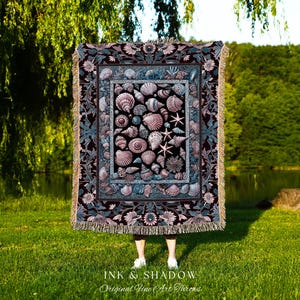 May include: A rectangular throw blanket featuring a detailed seashell design in shades of black, teal, and pink. The blanket has a floral border and fringe trim. The text "INK & SHADOW Original Fine Art Throws" is printed at the bottom.