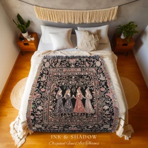 May include: A woven throw blanket featuring a black background with a central illustration of four figures in long dresses and pointed hats, surrounded by a floral border. The text "INK & SHADOW Original Fine Art Throws" is printed at the bottom.