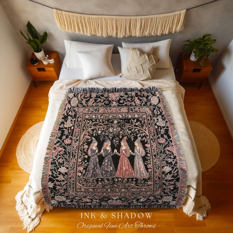 May include: A woven throw blanket featuring a black background with a central illustration of four figures in long dresses and pointed hats, surrounded by a floral border. The text "INK & SHADOW Original Fine Art Throws" is printed at the bottom.