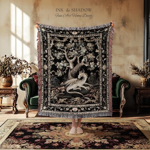 May include: A woven throw blanket featuring a central illustration of a white dragon resting beneath a large tree on a black background. The blanket has a decorative floral border and fringed edges. The text INK & SHADOW Fine Art Home Decor is visible.