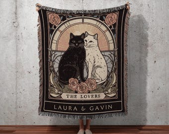 May include: A black and cream-colored throw blanket featuring a graphic of two cats, roses, and the words "The Lovers" and "Laura & Gavin." The blanket has a fringed edge and the text "INK & SHADOW Original Fine Art Throws."