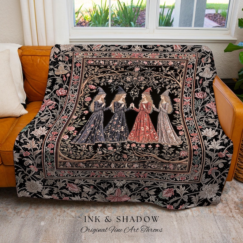 May include: A woven throw blanket featuring a black background with a central rectangular panel depicting four figures in pointed hats and long dresses standing among stylized trees and flowers. The border displays floral patterns in pink and white.