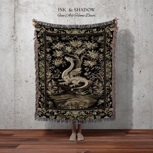 May include: A woven throw blanket featuring a central illustration of a light-colored dragon perched beneath a flowering tree on a black background. The design is framed by a decorative floral border with fringed edges and the text INK & SHADOW.