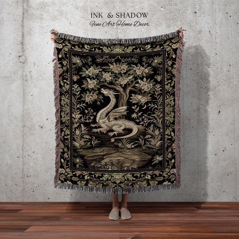 May include: A woven throw blanket featuring a central illustration of a light-colored dragon perched beneath a flowering tree on a black background. The design is framed by a decorative floral border with fringed edges and the text INK & SHADOW.