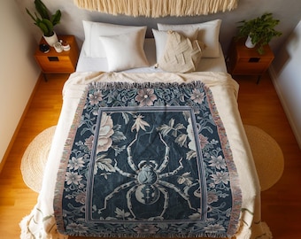 May include: A dark blue throw blanket with a large spider design, surrounded by floral patterns in pink, white, and green. The blanket has a fringed edge and the text "INK & SHADOW Original Fine Art Throws".