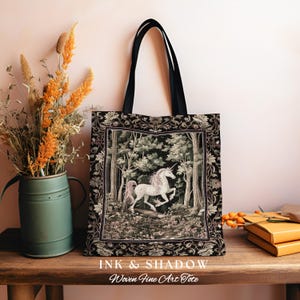 Może przedstawiać: Czarna torba typu tote z tkaniny z motywem gobelinu przedstawiającym białego jednorożca w lesie. Torba ma czarne paski i napis "INK & SHADOW Woven Fine Art Tote" nadrukowany na dole.
