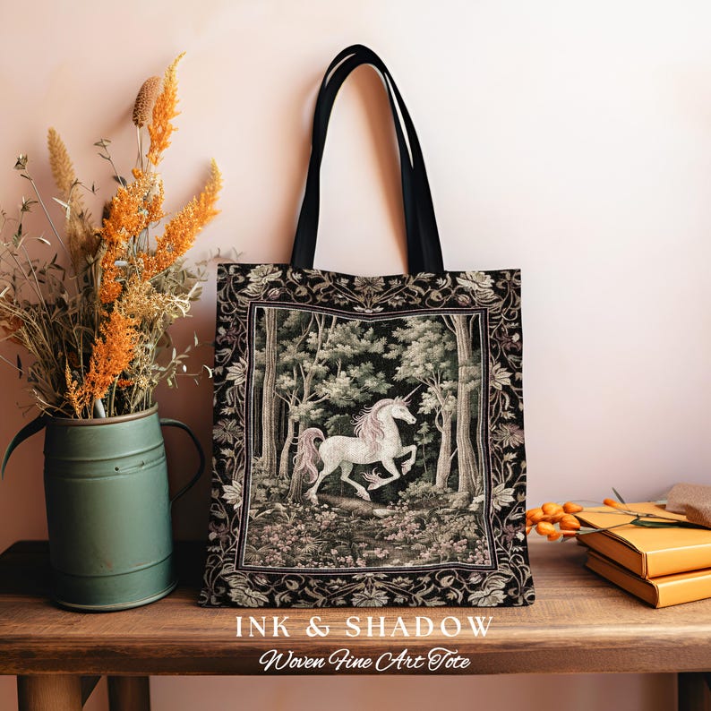 Może przedstawiać: Czarna torba typu tote z tkaniny z motywem gobelinu przedstawiającym białego jednorożca w lesie. Torba ma czarne paski i napis "INK & SHADOW Woven Fine Art Tote" nadrukowany na dole.
