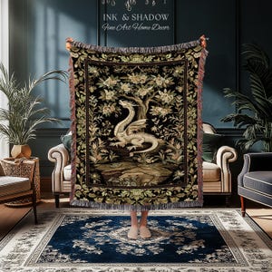 May include: A woven throw blanket featuring a central illustration of a white dragon perched on a rock beneath a flowering tree, set against a black background with a decorative floral border and fringed edges, displaying the text Ink & Shadow.