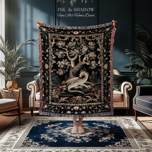 May include: A woven throw blanket featuring a central illustration of a white dragon resting beneath a large tree, surrounded by a dark floral border. The blanket has fringed edges and displays the text INK & SHADOW Fine Art Home Decor.