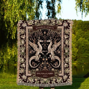 Könnte beinhalten: Eine weinrote Decke mit einem detaillierten Design mit zwei geflügelten Drachen, die sich gegenüberstehen. Die Decke hat einen floralen Rand und die Worte "Kindred Spirits" und "Dylan & Gabby 2024". Die Decke ist gefranst und trägt den Text "INK & SHADOW Original Fine Art Throws".