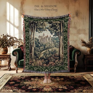 May include: A woven tapestry throw blanket featuring a detailed landscape scene of a castle nestled among trees. The blanket has a dark green border with floral patterns and fringed edges. Text at the top reads INK & SHADOW Fine Art Home Decor.