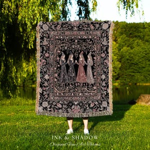 May include: A rectangular woven throw blanket featuring a black background with a central illustration of four figures in pointed hats and long dresses standing in a circle, surrounded by a detailed floral border and fringed edges.