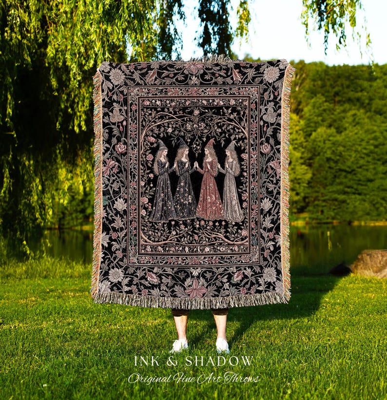 May include: A rectangular woven throw blanket featuring a black background with a central illustration of four figures in pointed hats and long dresses standing in a circle, surrounded by a detailed floral border and fringed edges.