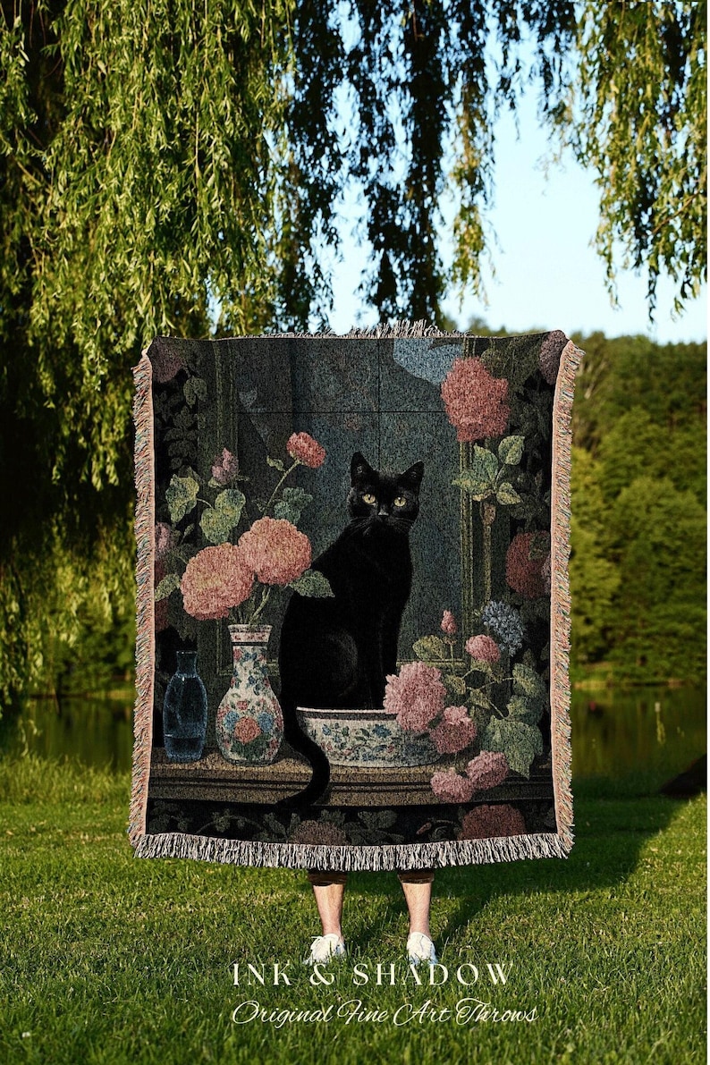 May include: A tapestry throw blanket featuring a black cat sitting amidst floral arrangements in a window. The throw has a dark background with pink and blue flowers, vases, and a decorative border. The text "INK & SHADOW Original Fine Art Throws" is at the bottom.