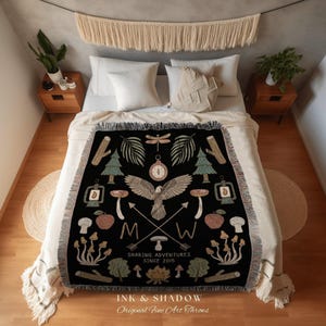 May include: Black throw blanket with a nature-themed design. Features an eagle, compass, trees, mushrooms, and the text "SHARING ADVENTURES SINCE 2015". The blanket is fringed and has the text "INK & SHADOW Original Fine Art Throws".