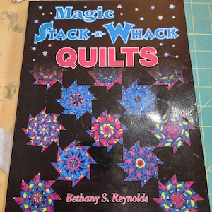 May include: A softcover instructional book titled Magic Stack-n-Whack QUILTS by Bethany S. Reynolds. The black cover features a repeating pattern of colorful, star-shaped quilt blocks in shades of blue, pink, and yellow, accented by small stars.
