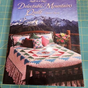 May include: A softcover instructional book titled Quilt in a Day Delectable Mountains Quilt by Eleanor Burns. The cover features a photograph of a bed with a pink and blue patterned quilt, several decorative pillows, and a mountain landscape.