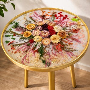 Op de afbeelding: Een ronde bijzettafel met een goudkleurig metalen frame en drie poten. Het tafelblad heeft een helder hars oppervlak waarin een levendig arrangement van gedroogde bloemen in tinten geel, roze, rood en groen is verwerkt, met gouden accenten.