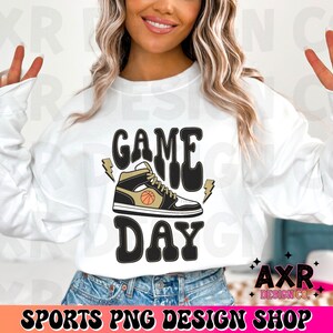 May include: White sweatshirt with the text "GAME DAY" in black and gold lettering. A black and gold basketball shoe with a basketball graphic is above the text.