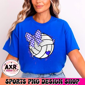 Op de afbeelding: Een blauw T-shirt met een witte volleybal met een paars-wit geruit strikje erop. De volleybal heeft een paars hartje en is omringd door paarse glitter.